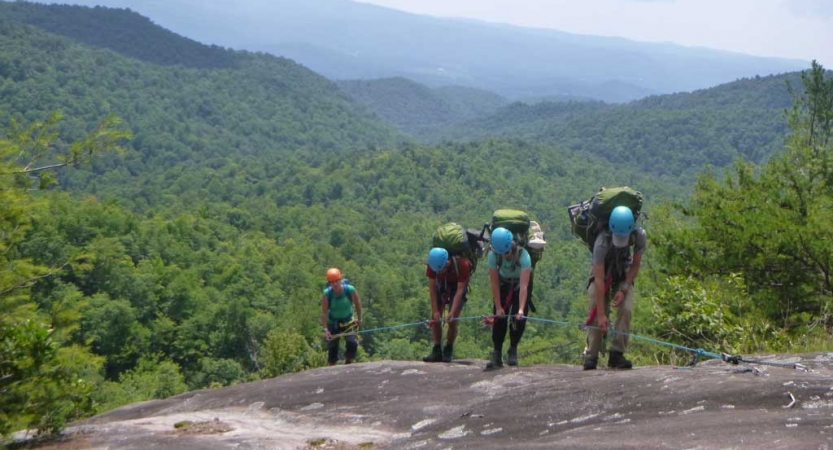 Four people wearing safety gear hold onto a rope as they hike along a rocky incline. The blue ridge mountains are in the background. 